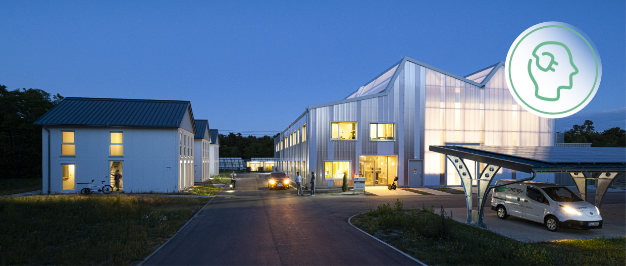 die SEnSSiCC-Halle bei Anbruch der Dunkelheit. Halbtransparente Fassade hell erleuchtet. Links daneben das Living Lab, rechts im Vordergrund ein Carport.