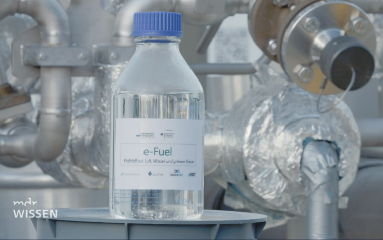 Glasflasche mit durchsichtigem synthetischen Kraftstoff (E-Fuels) steht vor den Rohren der Power-to-X-Anlage. Damit gelingt die Verkehrswende, Energiewende