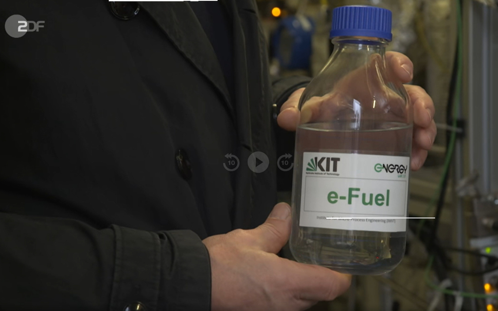 Professor Dittmeyer zeigt eine Glasflasche mit synthetischem Kraftstoff.. Der Flascheninhalt - E-Fuel - ist transparent.